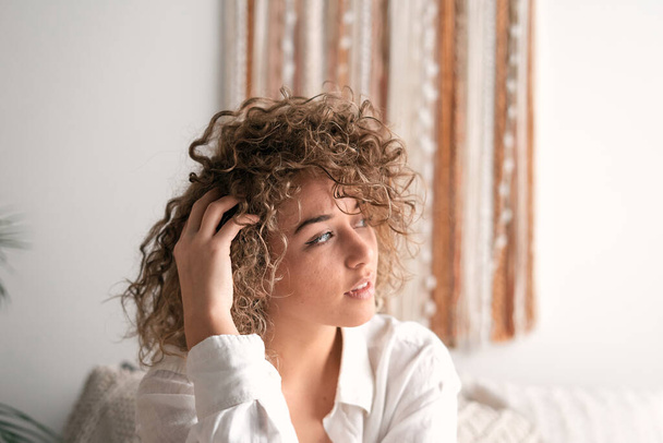 Jeune femme en chemise blanche touchant les cheveux bouclés et regardant loin dans la chambre à coucher lumineuse le matin - Photo, image