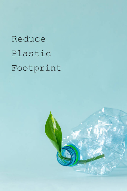 Murskattu muovipullo, jossa nuori vihreä lehti kasvaa siitä, Kirjoitus vähentää muovijalanjälkeä, Ympäristökonsepti, Pastellin sininen tausta, Pystysuora valokuva - Valokuva, kuva