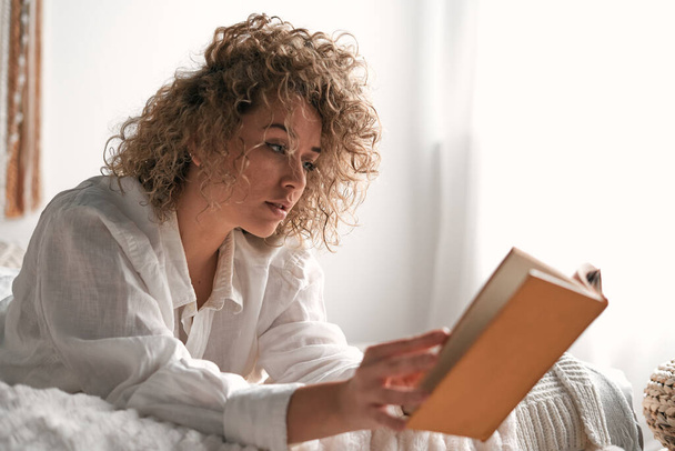 Ruhige junge Frau mit lockigem blondem Haar in lässiger Kleidung liegt auf einem bequemen Bett und liest an einem sonnigen Morgen im gemütlichen Schlafzimmer interessante Bücher - Foto, Bild