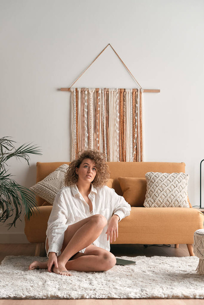 Mujer joven en camisa blanca tocando el pie y mirando hacia otro lado mientras está sentada en una alfombra suave cerca del sofá con cojines en la sala de estar iluminada por el sol en casa - Foto, Imagen
