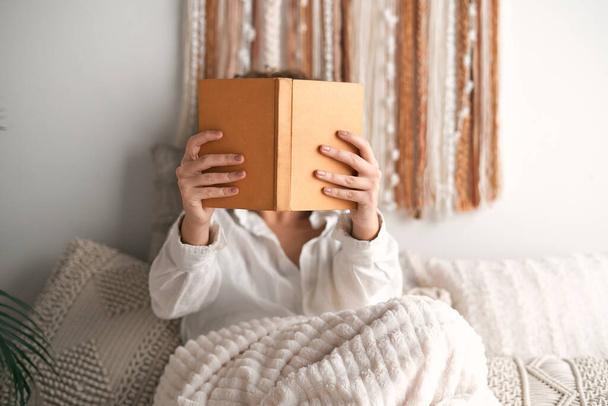 Unrecognizable female in white shirt sitting on bed under blanket and reading book in outstretched arms in light bedroom in weekend at home - Photo, image