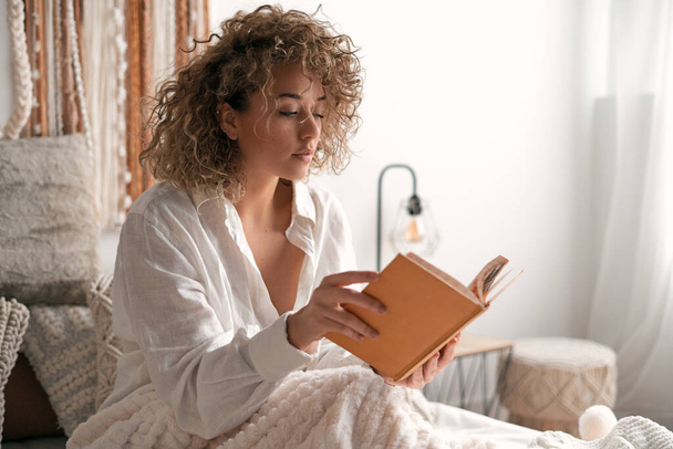 Młoda kobieta w białej koszuli z kręconymi włosami siedzi na miękkim łóżku i cieszy się czytaniem w słonecznej sypialni w weekendowy poranek w domu - Zdjęcie, obraz