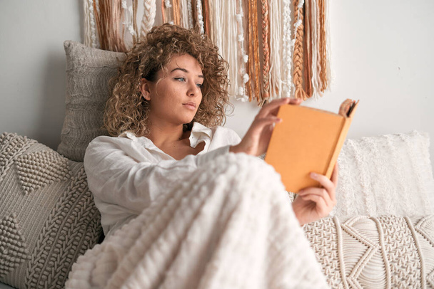 Молодая женщина в белой рубашке с вьющимися волосами сидит возле мягких подушек и читает интересную книгу в выходные утром в уютной спальне дома - Фото, изображение