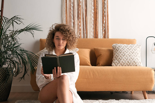 Потрясающая молодая женщина в повседневной одежде, сидящая на полу возле дивана и читающая книгу, проводя выходные дома - Фото, изображение