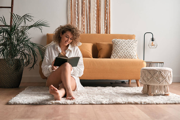 Full body of barefoot female sitting on floor and reading interesting book while chilling cozy living room on weekend - Фото, изображение