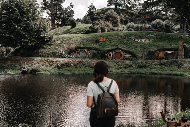 caucasian girl from the back dressed in a white t-shirt, a backpack and pants contemplating calmly and relaxed the house that is located inside the mountain on the other side of the lake surrounded by - Photo, Image