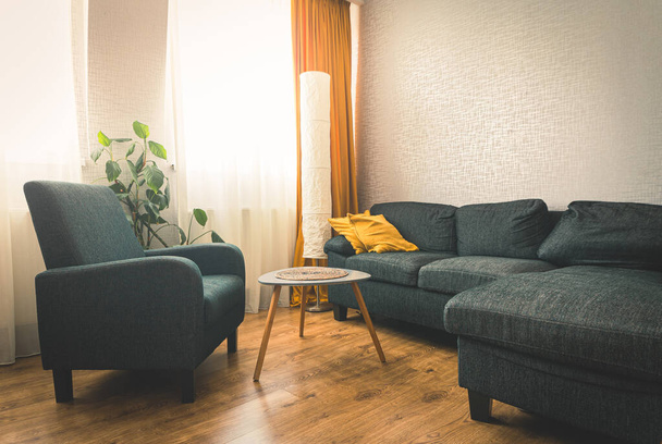 Hangulatos apartman nappali kanapéval és sárga párnák és elegáns asztal könyv virággal és ablakkal kék fény kívül télen. Szürke karosszék.másolás paszta fal háttér - Fotó, kép