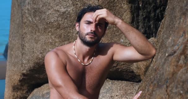 Attractive bare-chested macho man touches hair fixes hairstyle, mysteriously looking at camera, rests on the beach rocks - Footage, Video