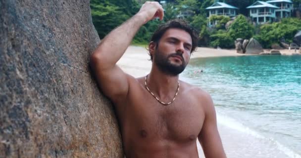 Aantrekkelijke jonge macho man, staat bloot-borst op het strand, dromerig opzij kijken dan zelfverzekerd staren naar de camera - Video