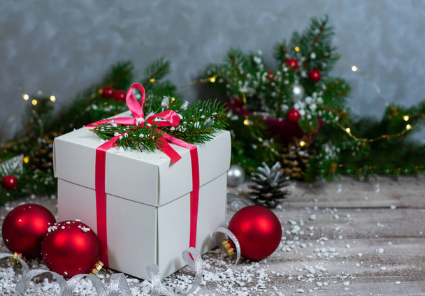 Una foto de una caja de regalo blanca que se encuentra sobre una mesa de madera entre los juguetes del árbol de Navidad y las ramas de coníferas contra una pared con una textura abstracta de color gris. - Foto, imagen