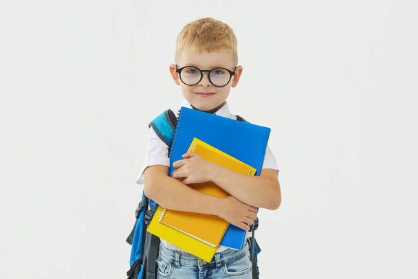 Back to school. Funny little boy from elementary school with a book, a backpack and glasses on a white background. Laughs merrily. Preparation for school. Concept of early development - Foto, imagen