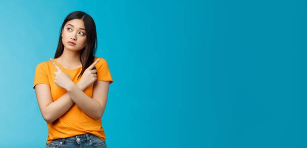 Confused cute asian female student thinking what do, look sideways thoughtful, cross hands chest pointing left and right, making choice, thinking what pick, taking important decision blue background. - 写真・画像