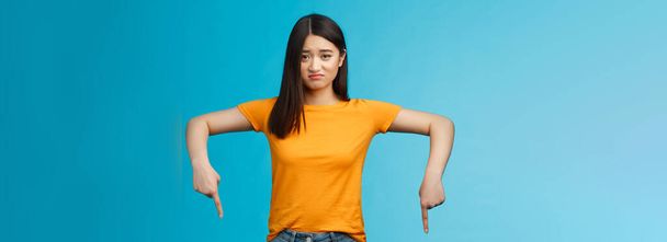 Moody sad asian brunette girl in yellow t-shirt smirking unsatisfied frowning bothered, complaining pointing down index fingers, showing bottom advertisement upset, jealous feel regret. - Fotografie, Obrázek