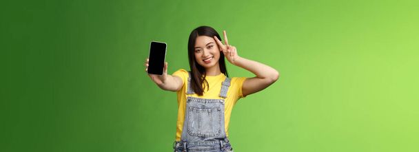 Cute beztroski azjatycki brunetka pokazując aplikację na ekranie smartfona, zrobić znak pokoju zwycięstwo, uśmiechając się radośnie, chwalić popularność mediów społecznościowych, ilość zwolenników, stać zielone tło. - Zdjęcie, obraz