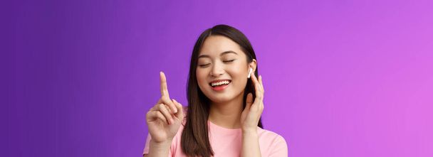Közelkép vidám boldog vonzó ázsiai nő csukott szemmel felemeli az egyik ujját vár kórus, mosolygós örömteli érintés vezeték nélküli fülhallgató, hogy hangerő hangosabb elégedett élvezze dalok, lila háttér. - Fotó, kép
