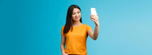 Elegante chica linda asiática guapa sostiene teléfono inteligente hablando video-llamada sonriendo amplia pantalla del teléfono, tomando selfie, sintiéndose bonita, hablando seguidores en línea medios sociales, fondo azul. - Foto, imagen