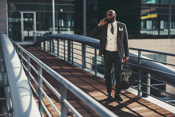 A mature fashionable black man entrepreneur is talking on the phone while passing on an overhead passage; an African bearded businessman is phoning while standing on the pedestrian bridge with his bag - Foto, immagini