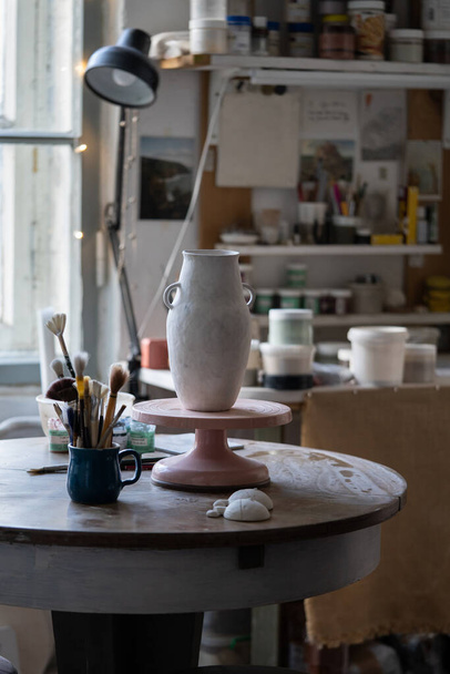 Clay vase dries up standing on wooden rack. Masterpiece prepared for making sketch and painting stands on table against tools for sculpturing. Handmade vase created for sending to exhibition in museum - 写真・画像