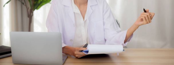 Young woman doctor using laptop computer and looking document paper about patient for diagnostic at clinic, physician female working and reading document on clipboard, one person, medical concept. - Foto, Imagem