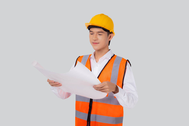 Portrait young asian man is engineer wearing helmet holding and looking blueprint with confident isolated on white background, handsome male is architect or contractor, worker or labor concept. - 写真・画像