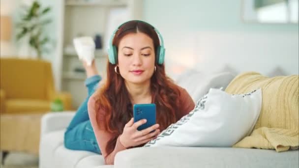 Relax, music and phone with woman in living room and headphones listening to radio, podcast or podcast. Happy, freedom and internet with girl on sofa, streaming for audio, social media or online. - Footage, Video