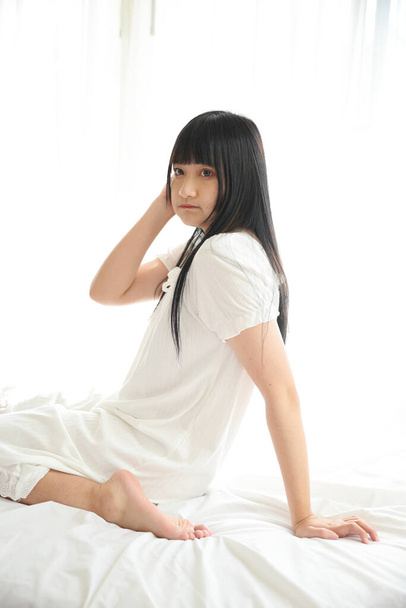 Hermosa joven asiática sentada en la cama relajándose en la luz de la mañana con habitación blanca - Foto, Imagen