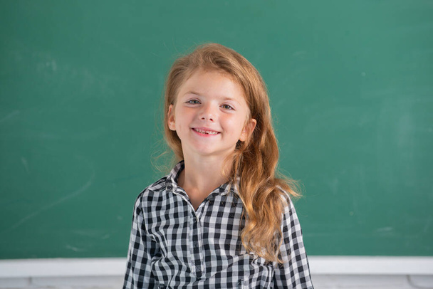 Close-up portrait of attractive small little cheerful girl sitting on table desktop in class room indoors. Little funny school girl face. Kids education and knowledge. Student kids - Фото, изображение