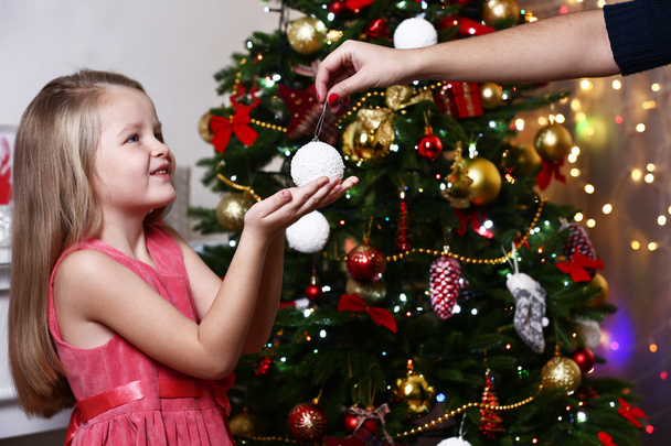 Little girl taking bauble near Christmas tree on white wall background - Foto, afbeelding