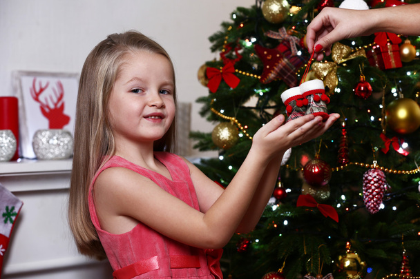 Little girl taking red little shoes near Christmas tree on white wall background - Foto, immagini