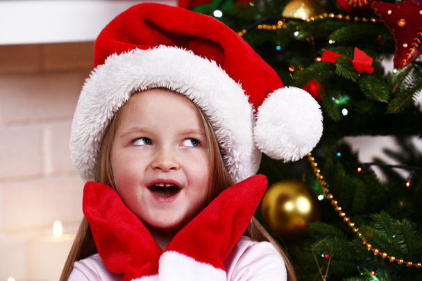 Little girl in Santa hat and mittens sitting near fir tree on fireplace with candles background - Fotó, kép