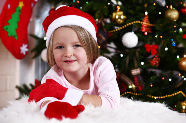 Little girl in Santa hat and mittens lying on fur carpet on Christmas tree background - Photo, Image