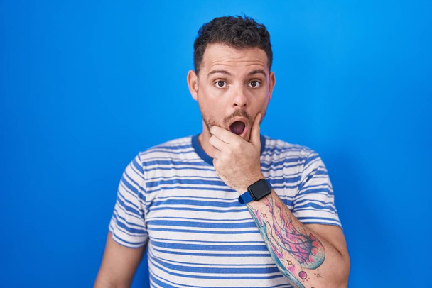 Young hispanic man standing over blue background looking fascinated with disbelief, surprise and amazed expression with hands on chin  - Photo, Image