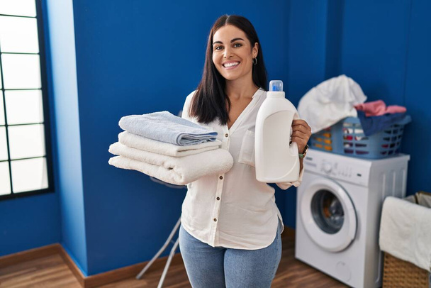 Mujer hispana joven sosteniendo ropa limpia y botella de detergente sonriendo con una sonrisa alegre y fresca en la cara. mostrando dientes.  - Foto, Imagen