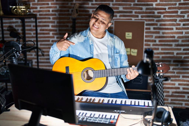 Spanyol fiatalember klasszikus gitáron játszik a zenei stúdióban, magabiztosan néz ki mosollyal az arcán, ujjaival büszkén és boldogan mutogat magára..  - Fotó, kép