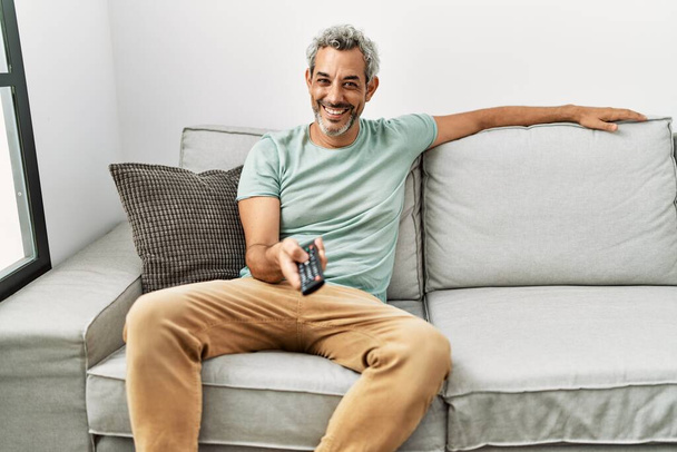 Седовласый мужчина средних лет смотрит кино, сидя дома на диване - Фото, изображение