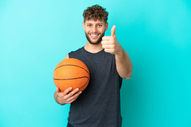 Schöner junger Mann spielt Basketball isoliert auf blauem Hintergrund mit erhobenem Daumen, weil etwas Gutes passiert ist - Foto, Bild