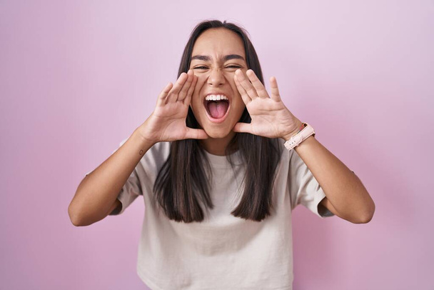 jonge latino vrouw staan over roze achtergrond schreeuwen boos hardop met handen over mond  - Foto, afbeelding