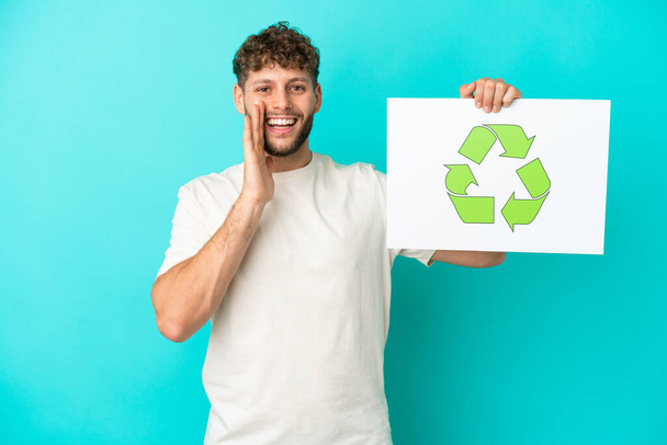 Junger gutaussehender kaukasischer Mann auf blauem Hintergrund, der ein Plakat mit Recycling-Symbol in der Hand hält und schreit - Foto, Bild