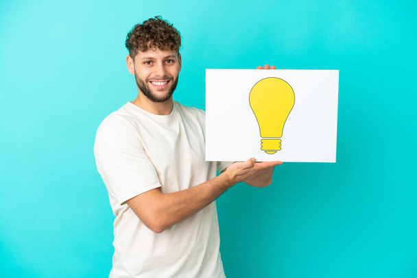 Jovem homem caucasiano bonito isolado no fundo azul segurando um cartaz com ícone de lâmpada com expressão feliz - Foto, Imagem