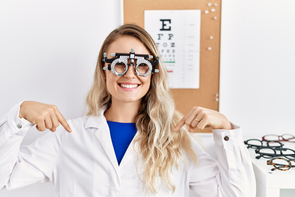 Junge schöne Optikerin mit Optometrie-Brille in der Klinik sieht selbstbewusst aus, mit einem Lächeln im Gesicht, und zeigt mit den Fingern stolz und glücklich.  - Foto, Bild