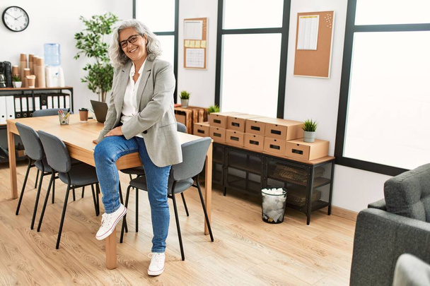 Middelbare leeftijd grijs-harige zakenvrouw glimlachen gelukkig zitten op de tafel op kantoor. - Foto, afbeelding