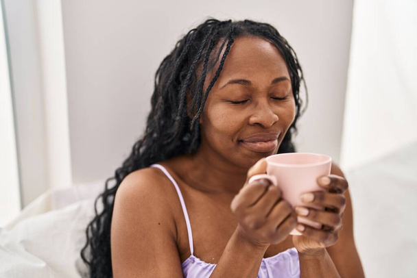 Afroamerykanka pije kawę siedząc na łóżku w sypialni - Zdjęcie, obraz