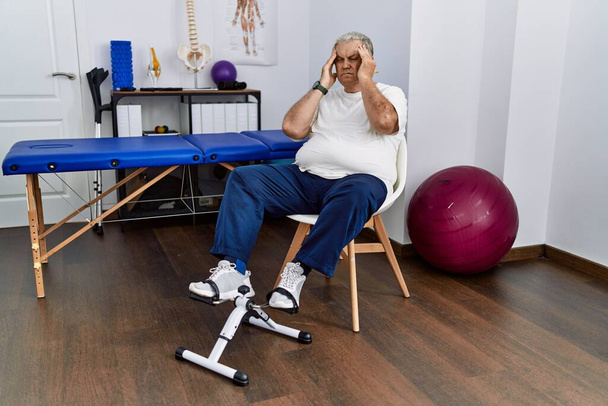 Senior caucasian man at physiotherapy clinic using pedal exerciser with hand on head for pain in head because stress. suffering migraine.  - Photo, Image