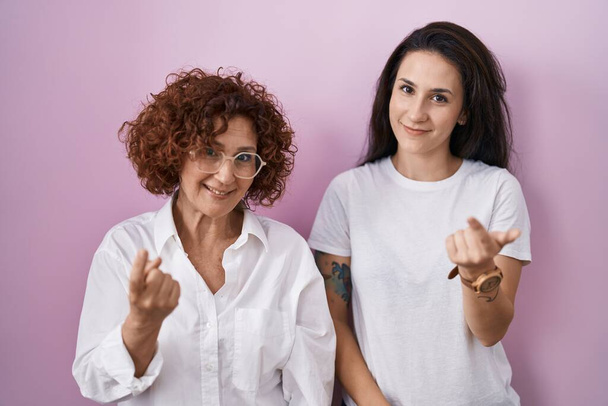 Madre e hija hispanas vistiendo camiseta blanca casual sobre fondo rosa haciendo señas vienen aquí gesto con la mano invitando a la bienvenida feliz y sonriente  - Foto, Imagen