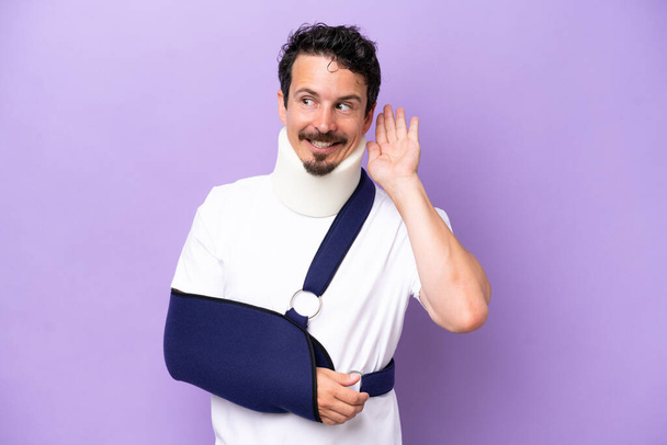 Young caucasian man wearing a sling and neck brace isolated on purple background listening to something by putting hand on the ear - Photo, Image