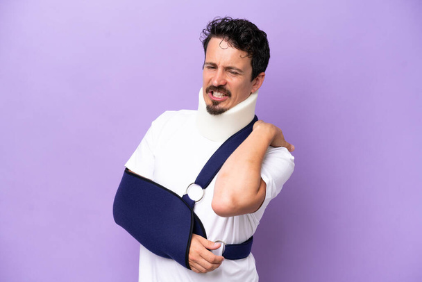 Junger kaukasischer Mann mit Tragetuch und Nackenstütze isoliert auf violettem Hintergrund, der unter Schulterschmerzen leidet, weil er sich bemüht hat - Foto, Bild
