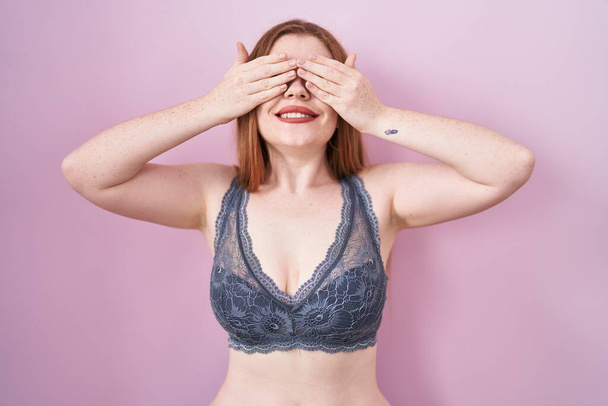 Redhead woman wearing lingerie over pink background covering eyes with hands smiling cheerful and funny. blind concept.  - Photo, Image