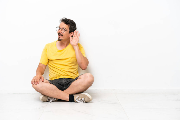 Fiatal kaukázusi férfi ül a padlón elszigetelt fehér háttér hallgat valamit azáltal, hogy kezét a fülre - Fotó, kép