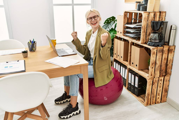 Beautiful caucasian business woman working at the office sitting on pilates ball screaming proud, celebrating victory and success very excited with raised arms  - Photo, Image