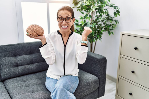 Young hispanic woman holding brain working at psychology clinic screaming proud, celebrating victory and success very excited with raised arms  - Foto, Bild